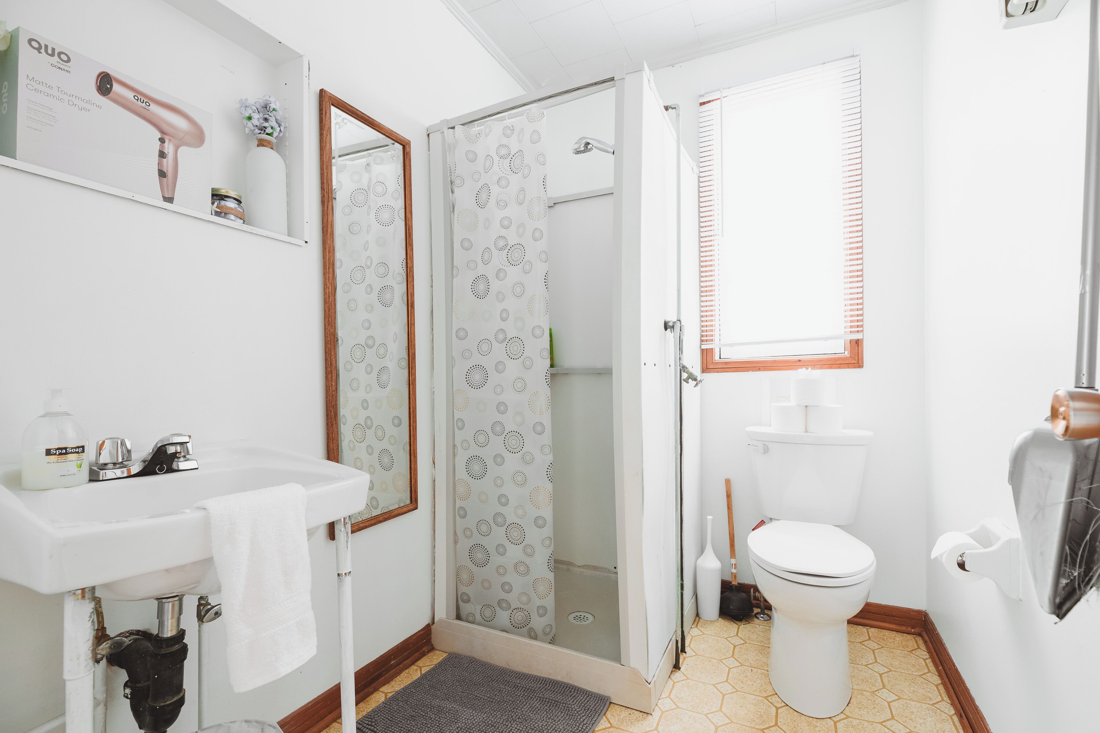 Une salle de bain avec un lavabo blanc, une douche avec des rideaux à motifs, des toilettes et une fenêtre avec des stores.