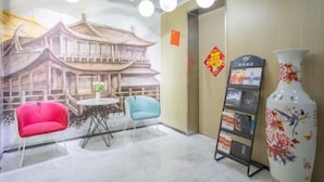 Ein Zimmer mit einer Wandmalerei traditioneller Architektur, einem roten Sessel, einem blauen Stuhl, einem kleinen Tisch, einer Ziervase und einem Broschürenständer.