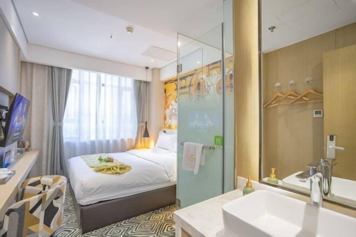 A hotel room with a bed, a desk with a computer, a large window with curtains, a bathroom with a sink and mirror, and a shower area.