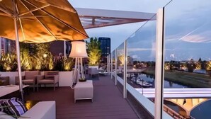 Ein moderner Außenbereich mit Sitzgelegenheiten, einem großen Sonnenschirm und Blick auf einen Fluss und Gebäude.