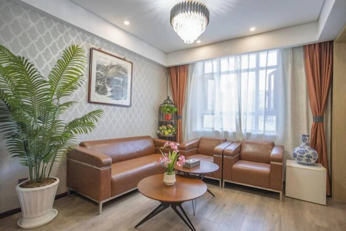 A living room with a sofa, a coffee table, a potted plant, a chandelier, and a window with curtains.