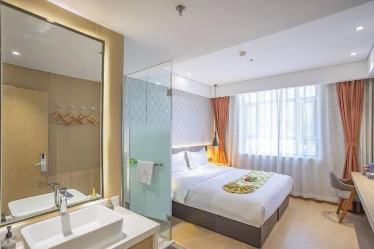 A hotel room with a large mirror, a bedside table, a bed, a desk, and a window with curtains.