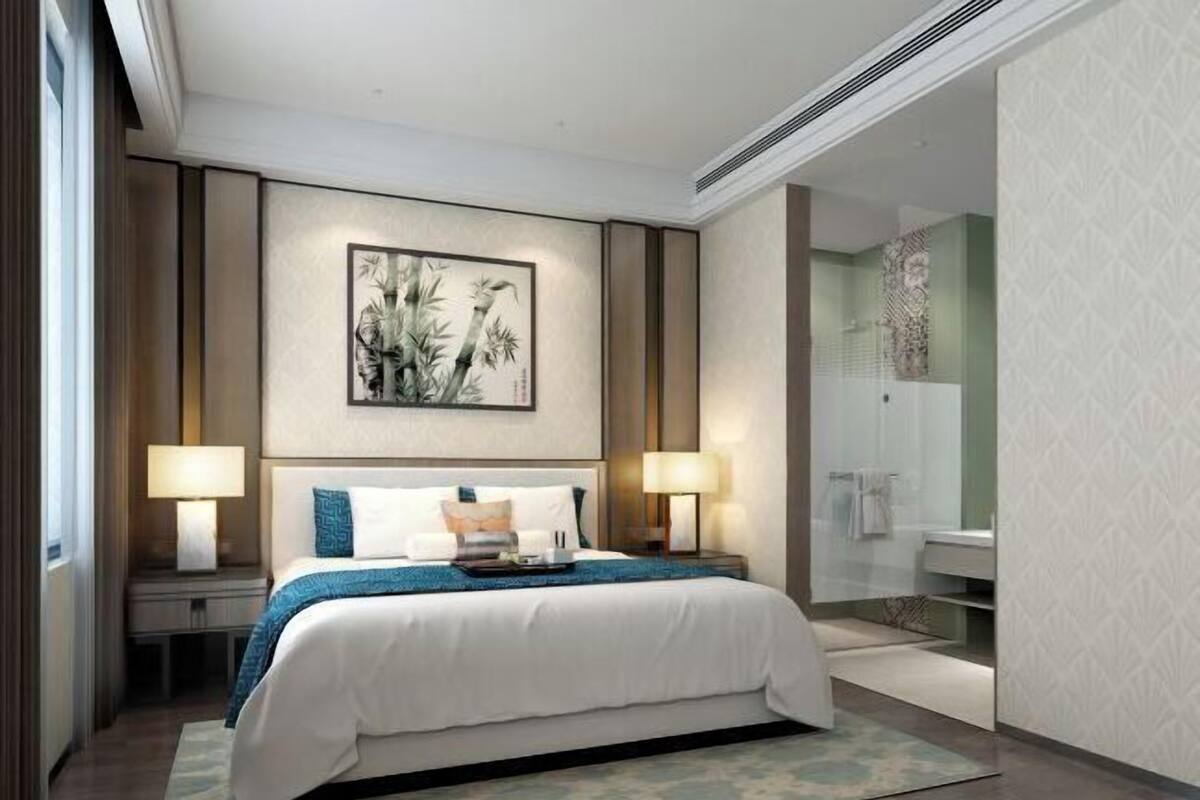 A modern hotel room with a large bed, two bedside lamps, a wall-mounted artwork, a bathroom with a glass shower, and a patterned wall.