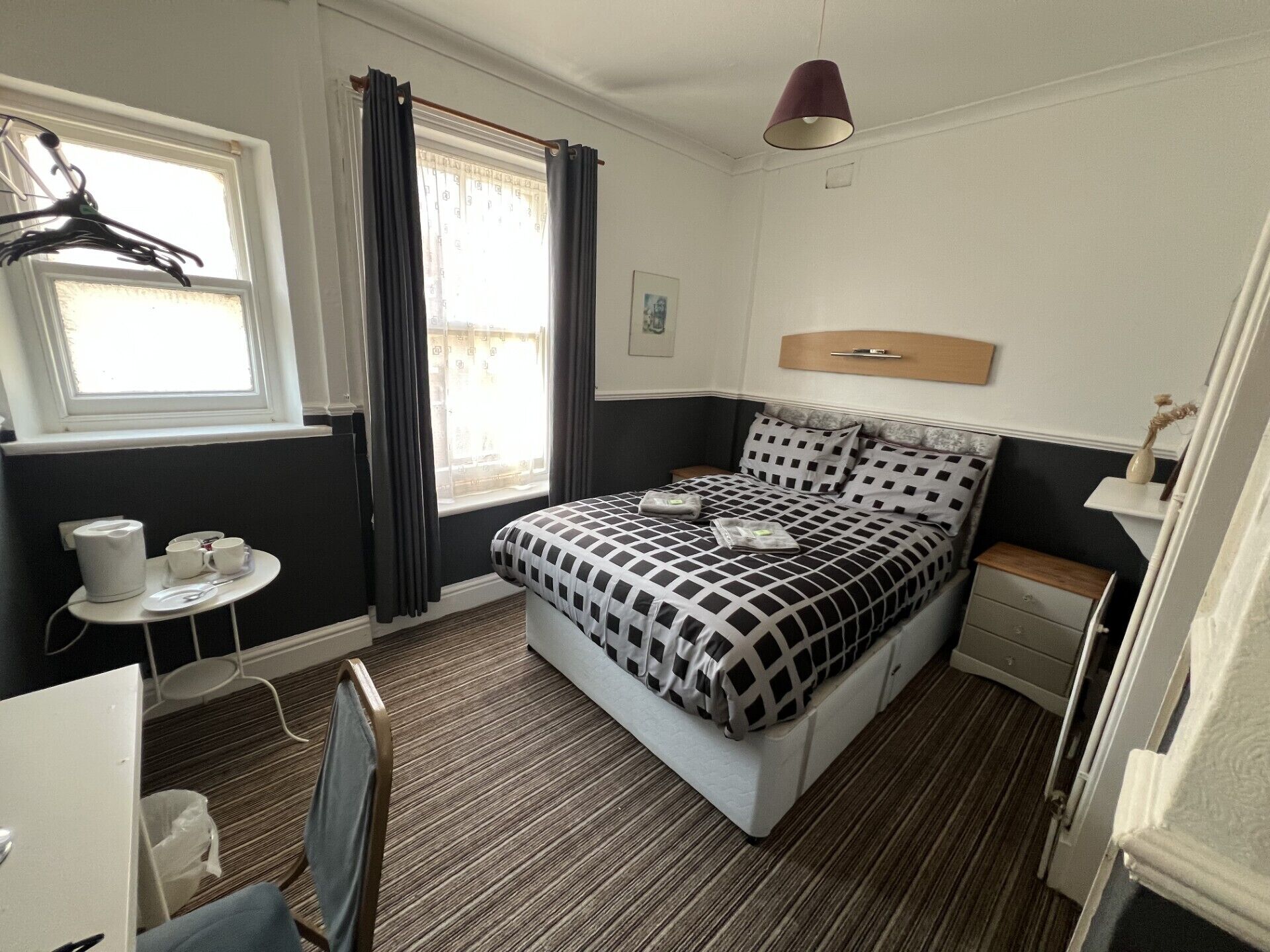 A hotel room with a bed, a bedside table, a chair, a small table with cups and a kettle, a window with curtains, and a wall-mounted lamp.