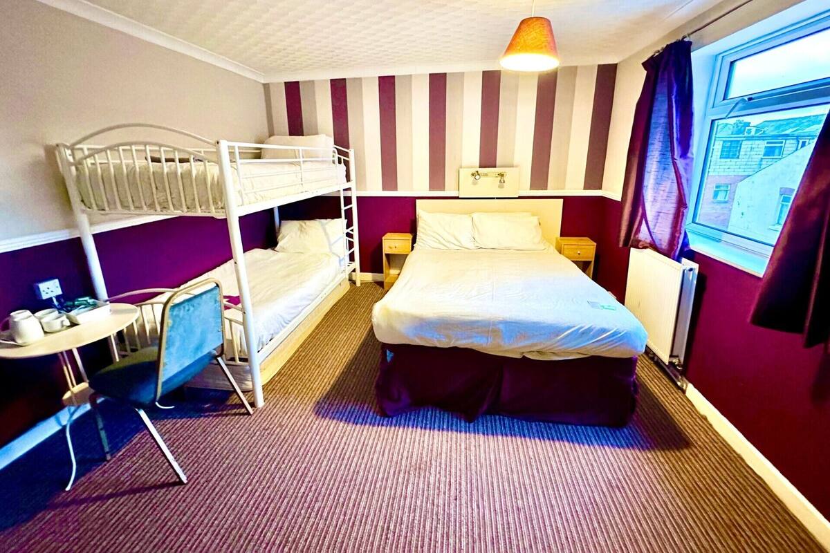 A hotel room with a bunk bed, a single bed, a desk with a chair, a lamp, a window with curtains, and a radiator.