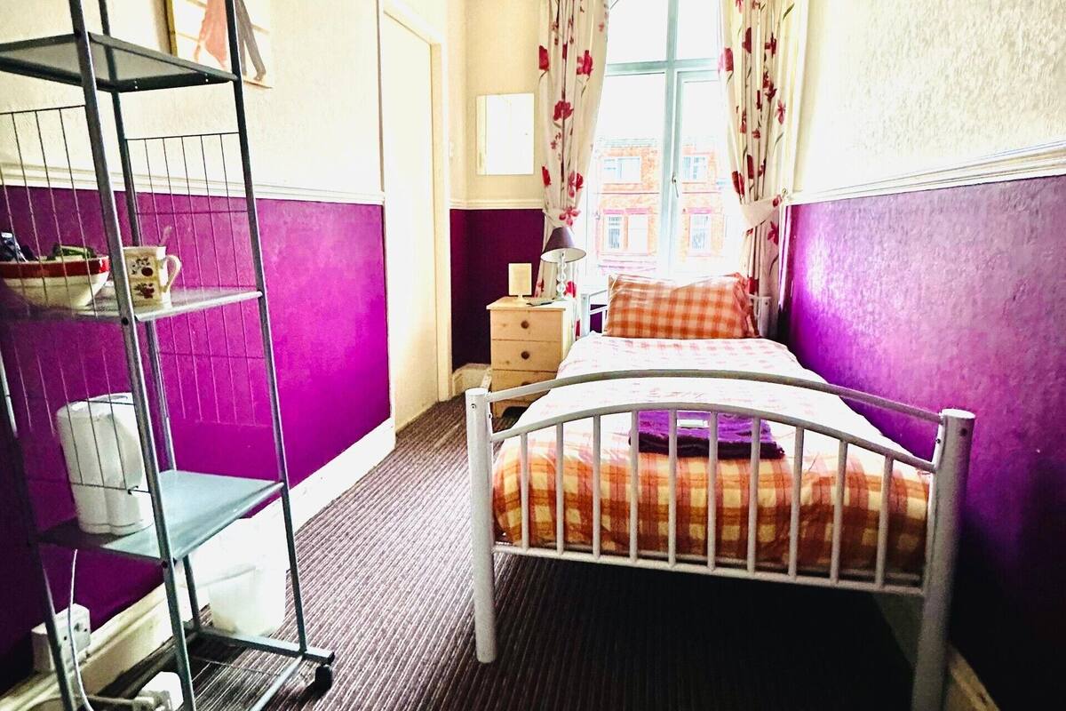 A compact room with a bed, a window with floral curtains, a metal shelf unit, and a ceiling light.