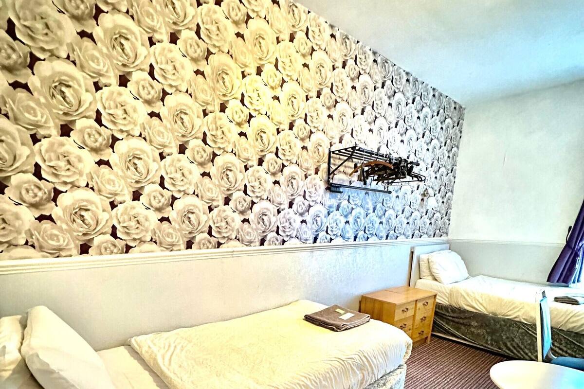 A hotel room with a large wall of artificial flowers, a sofa bed with white bedding, a small table, and a chair.