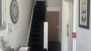 A hallway with a staircase leading to a private room. There is a clock on the wall, a door with a red handle, and a framed picture on the wall. - The AVON Hotel (Blackpool)