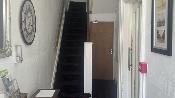 A hallway with a staircase leading to a private room. There is a clock on the wall, a door with a red handle, and a framed picture on the wall. - The AVON Hotel (Blackpool)
