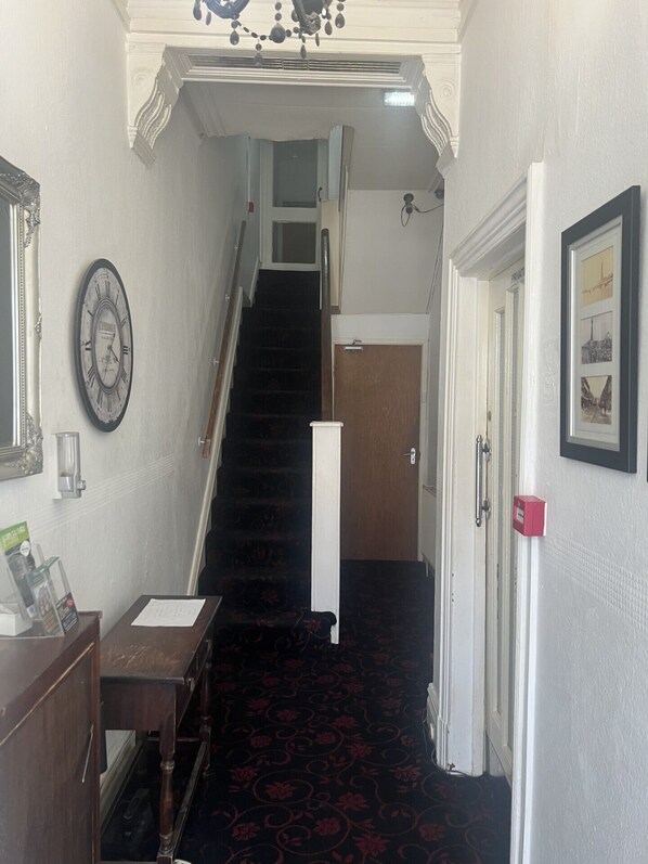A hallway with a staircase leading to a private room. There is a clock on the wall, a door with a red handle, and a framed picture on the wall. - The AVON Hotel (Blackpool)