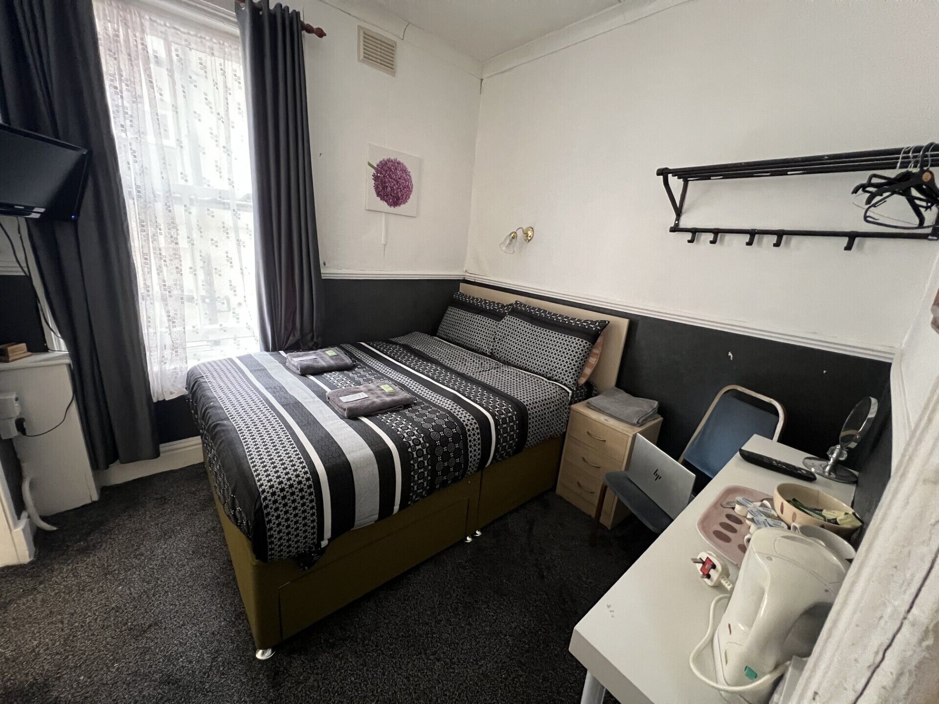 A compact hotel room with a single bed, a desk with a chair, a small bedside table, and a wall-mounted coat rack.
