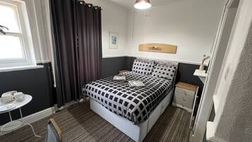 A compact hotel room with a bed, bedside table, and a small table with cups.