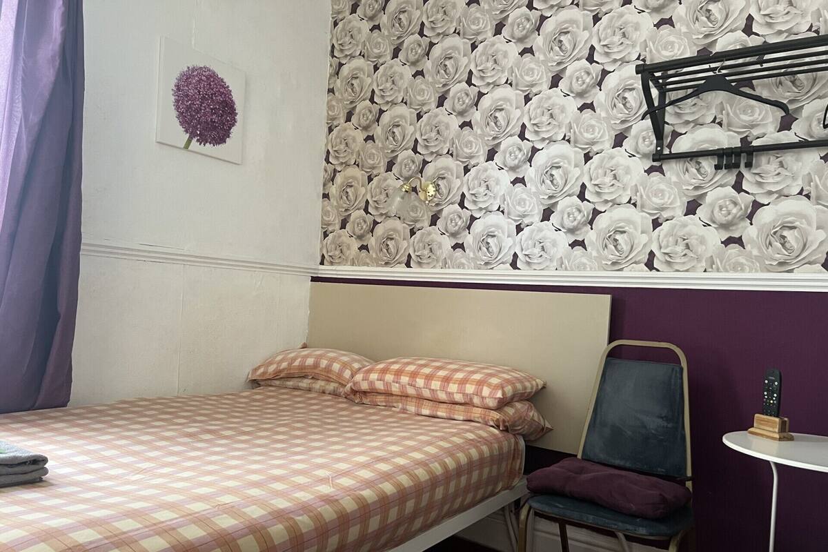 A bedroom with a bed, a chair, a small table, and a wall with a floral wallpaper.