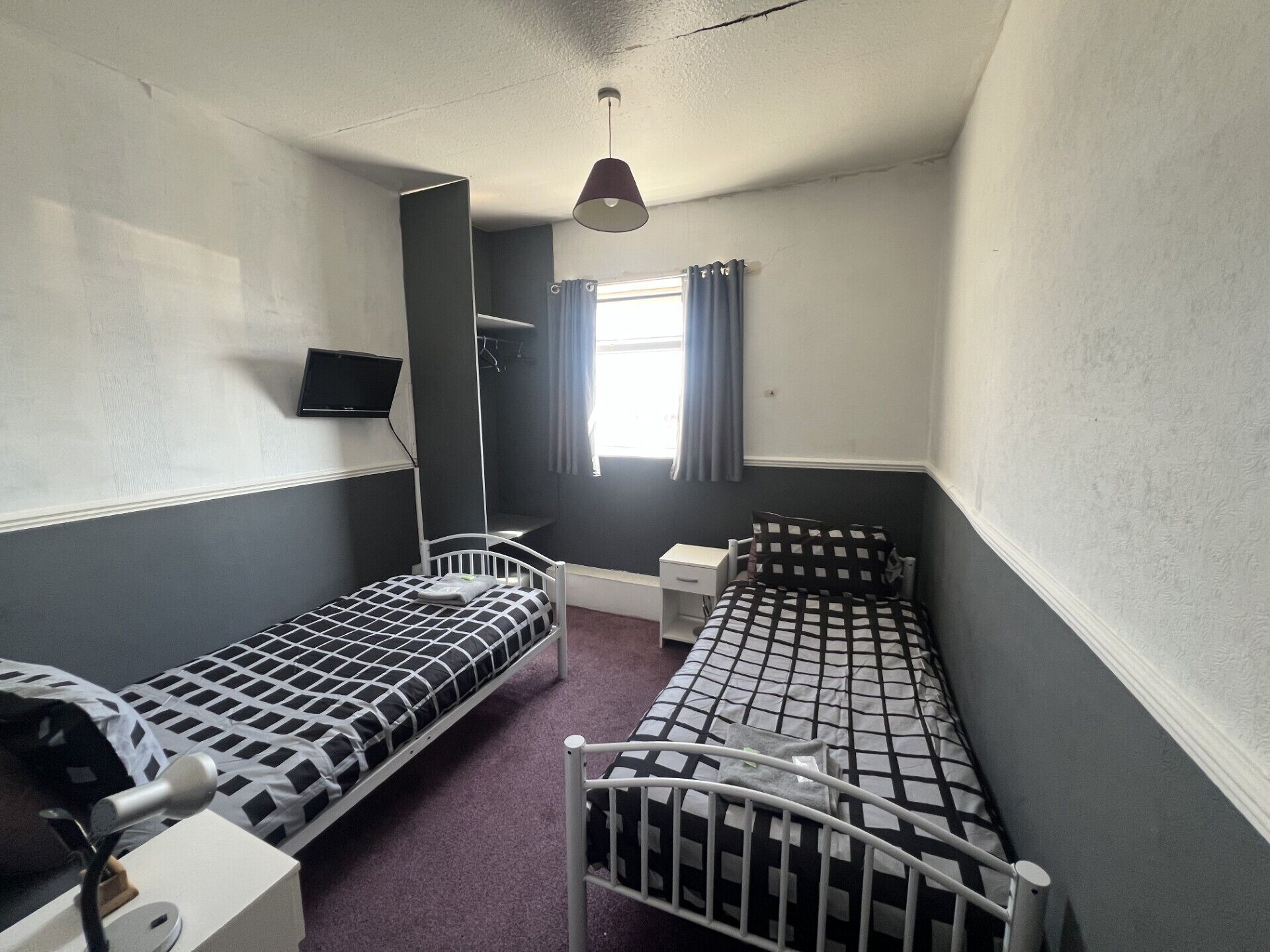 A small, single-room accommodation with two single beds, a bedside table, a chair, a desk, a flat-screen TV mounted on the wall, and a window with curtains.