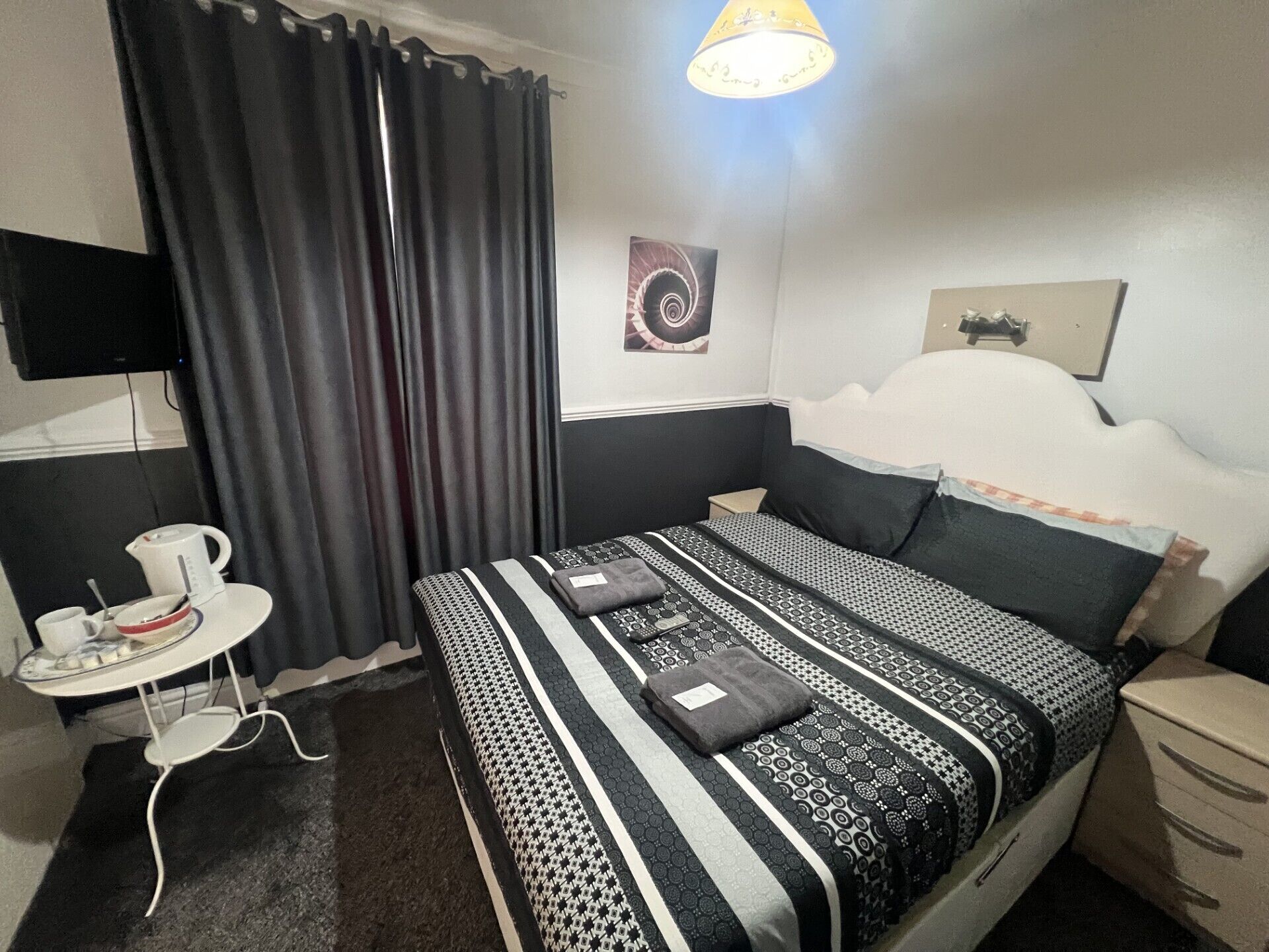 A neatly arranged hotel room with a bed, a small table with a kettle, and a wall-mounted TV.