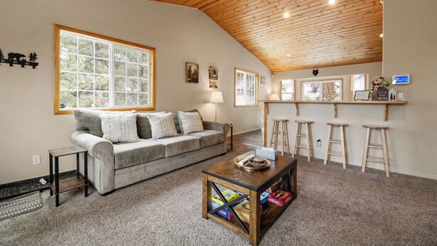 Una sala de estar acogedora con techo de madera, un sofá gris, una mesa de centro con libros y una zona de bar con taburetes.