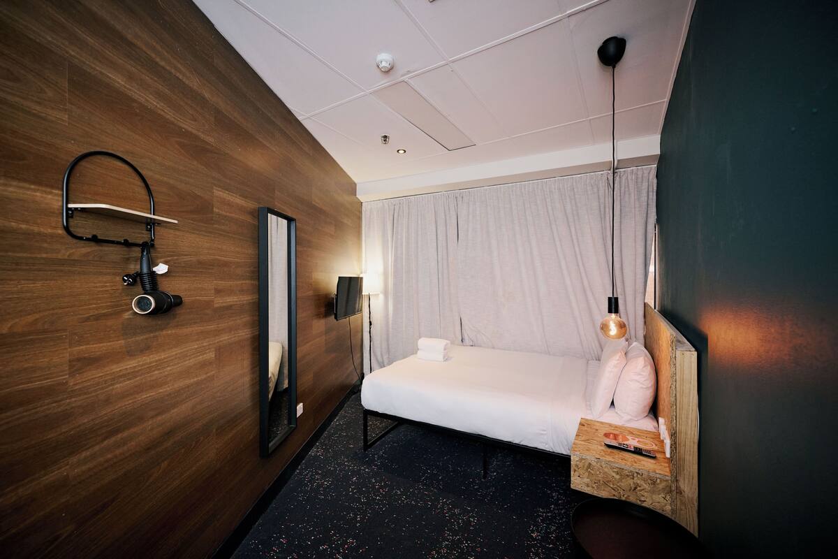 Habitación de hotel con pared de madera, cama con colchón blanco, mesita de noche con control remoto, espejo y lámpara colgante.