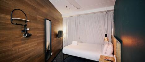 Habitación de hotel con pared de madera, cama con colchón blanco, mesita de noche con control remoto, espejo y lámpara colgante.