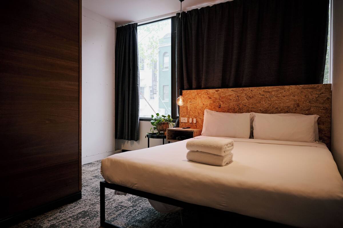 Habitación de hotel con cama, mesita de noche, planta en maceta y ventana con cortinas.