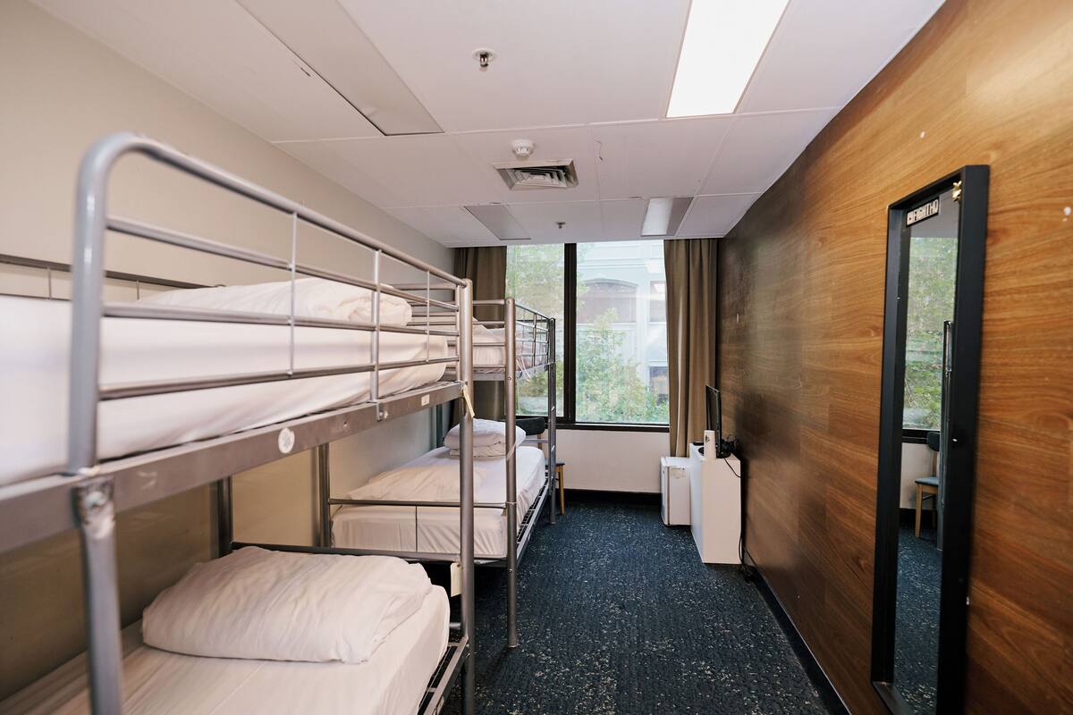 Dormitorio con literas metálicas, una mesita, una silla y una ventana con vistas a la vegetación.