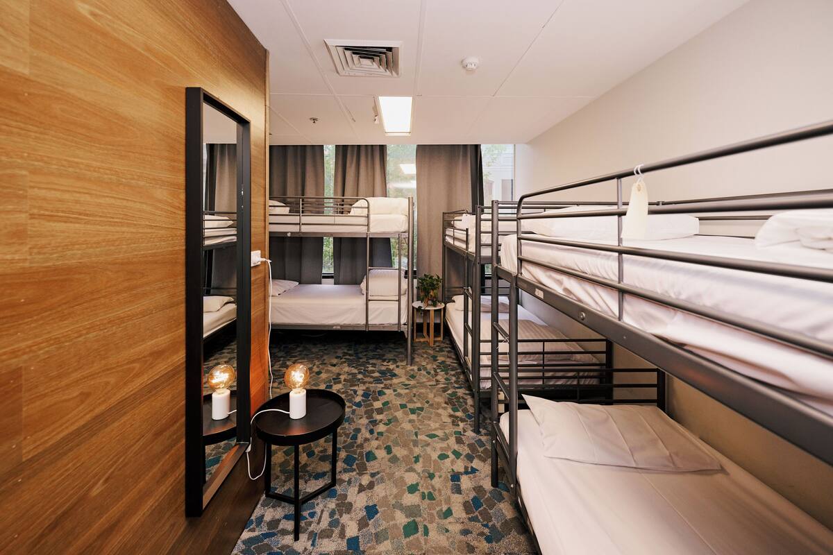 Una habitación de hotel con literas, un espejo, una mesita con una lámpara y una alfombra estampada.