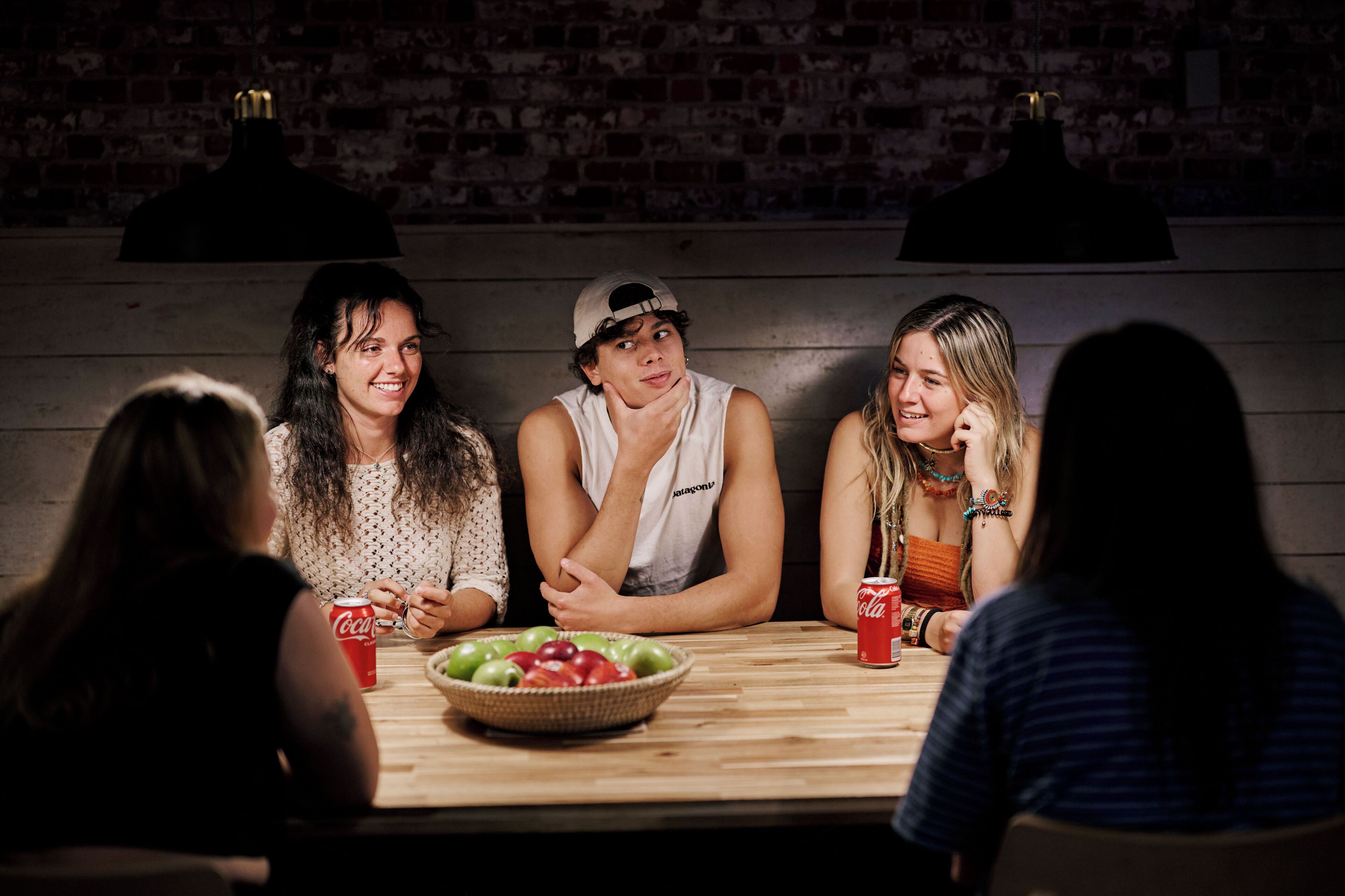 Un groupe de personnes est assis autour d’une table en bois, devant un saladier rempli de fruits et des boissons, dans une pièce aux murs de briques et éclairée par des lampes suspendues.