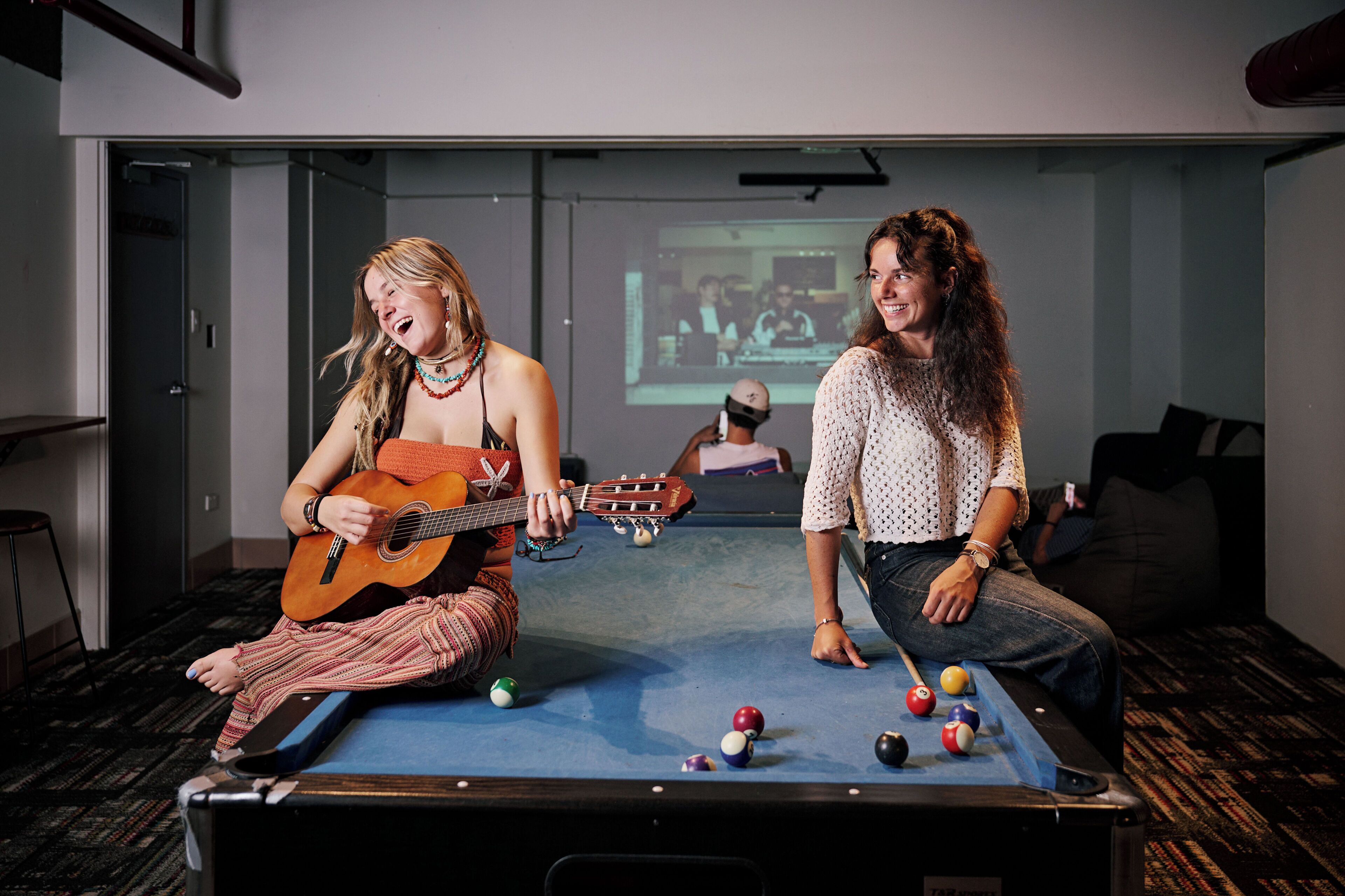 Dos personas jugando al billar y una tocando la guitarra en una habitación con pantalla de proyector.
