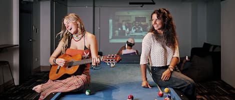 Dos personas jugando al billar y una tocando la guitarra en una habitación con pantalla de proyector.