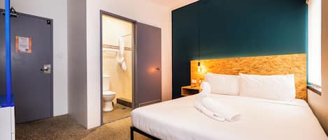 Habitación de hotel con cama, mesita de noche, cabecero de madera, baño con inodoro y lavamanos, y una puerta.