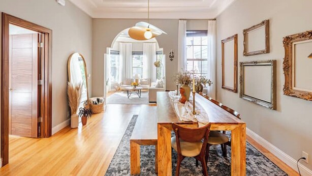 A spacious dining area with a long wooden table, a chandelier, and a view of a living room with a sofa and a window.