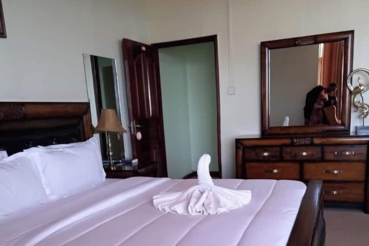 Una cama bien hecha con una toalla doblada encima, un aparador de madera con cajones, un espejo y una lámpara sobre una mesita de noche.