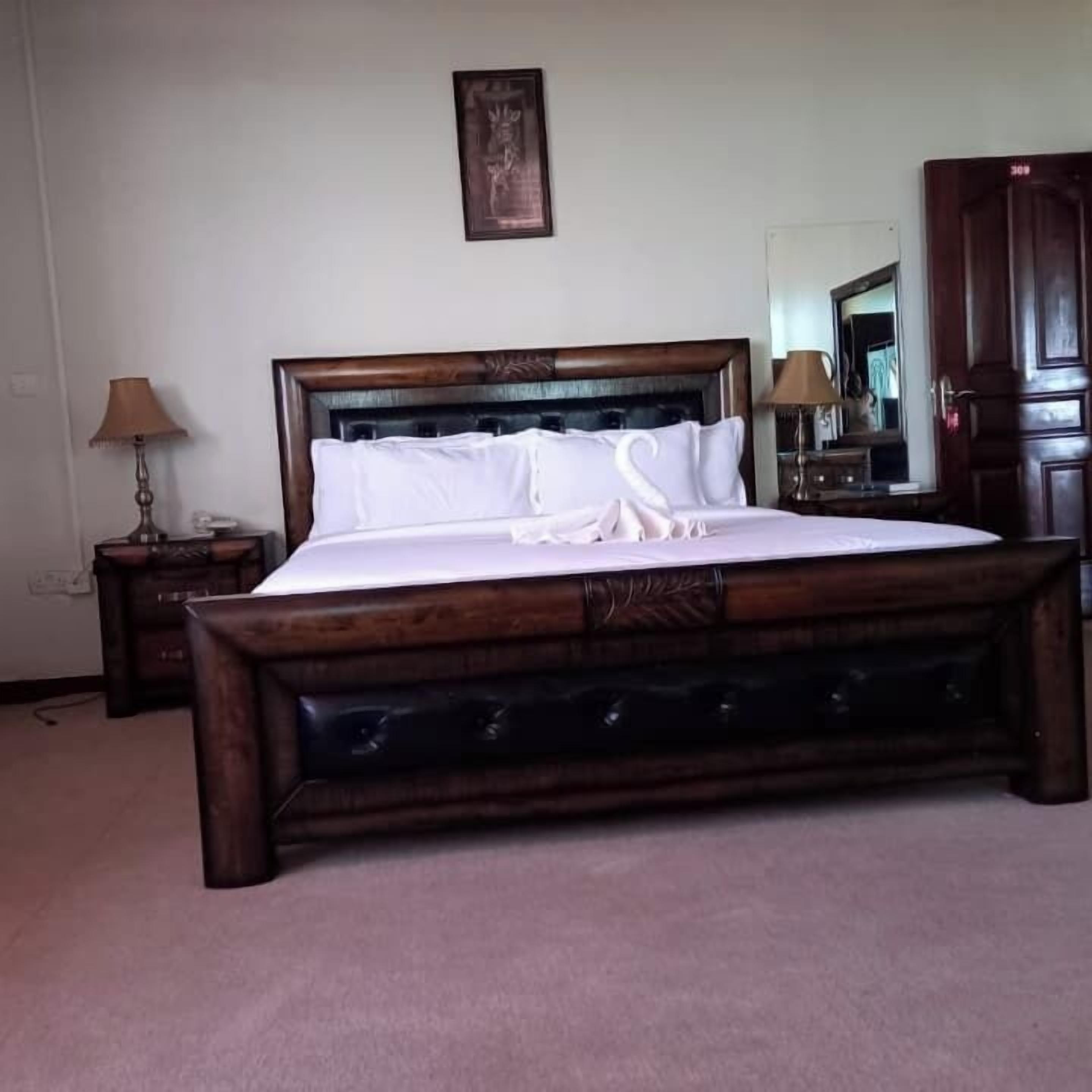 Un grand lit avec une tête de lit en bois, deux tables de chevet avec des lampes, un tableau encadré au mur et une armoire en bois.