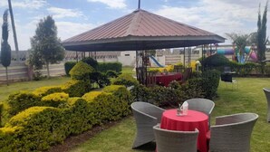 Un espace extérieur aménagé avec un coin salon abrité par une pergola, des fauteuils en osier et une petite table recouverte d’une nappe rouge.