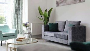 Una sala de estar con un sofá gris, una mesa de centro redonda de cristal, un sillón verde, una planta verde grande y un cuadro en la pared.