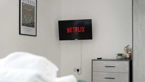 Un dormitorio con cama, cómoda, televisión mostrando 'NETFLIX' y un cuadro en la pared.