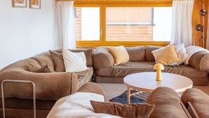 Ein gemütliches Wohnzimmer mit Holzbalkendecke, einem großen Fenster, einer Sofagarnitur und einem runden Tisch.