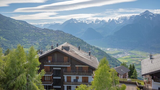 Eine Berghütte mit mehreren Balkonen, einer Holzfassade und Blick auf schneebedeckte Berge und ein Tal.
