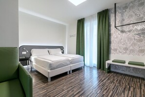 A modern hotel room with a bed, a green armchair, a bedside table with a lamp, and a wall-mounted shelf with green cushions. - Seele Garda Hotel (Nago-Torbole)