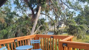 A wooden deck with blue folding chairs and a table, surrounded by lush greenery and a large tree. - Breezy Loft at Palmetto, FL. (Palmetto)