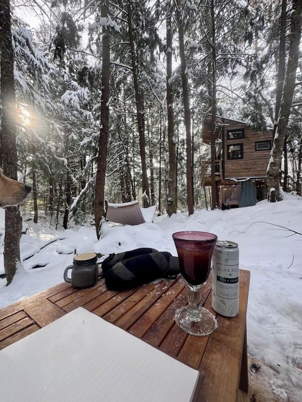A wooden table with a glass, a mug, and a can in a snowy forest setting. - Cozy Treehouse with Sauna in the Woods with Stream (Hardwick)