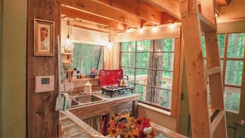 A rustic cabin interior with a wooden ceiling, a kitchen area with a red kettle, and a window overlooking a forest.