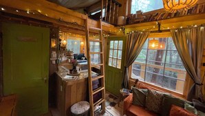 A cozy cabin interior with a wooden staircase, a kitchen area, a seating area with a sofa, and large windows with curtains. - Streamside treehouse in the Vermont forest with access to sauna (Hardwick)