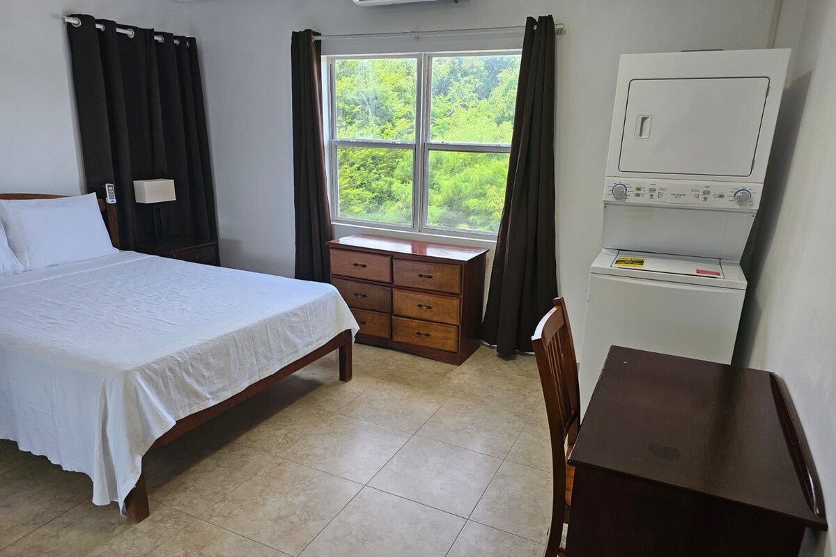 A hotel room with a bed, a wooden dresser, a desk, and a window with curtains.