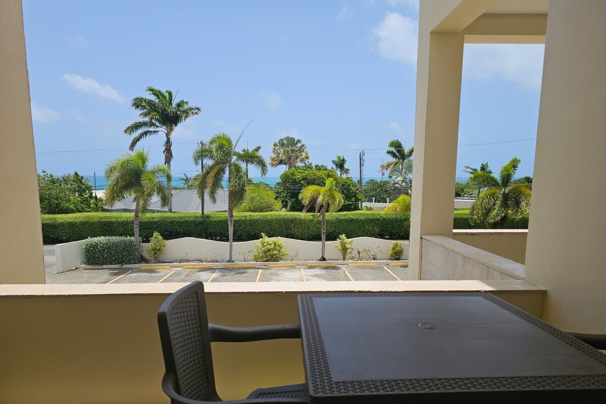 A balcony with a table and chair, overlooking a parking area and palm trees, with a clear view of the ocean.