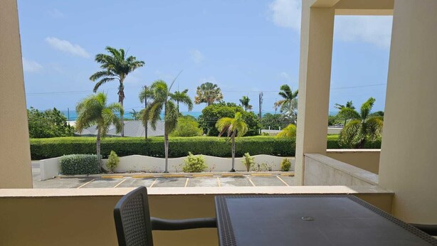 A balcony with a table and chair, overlooking a parking area and palm trees, with a clear view of the ocean. - Aurela Residences (St. John's)