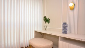 Esquina de una habitación con una cortina blanca, un taburete pequeño, una planta en un florero de vidrio y una lámpara de pared.
