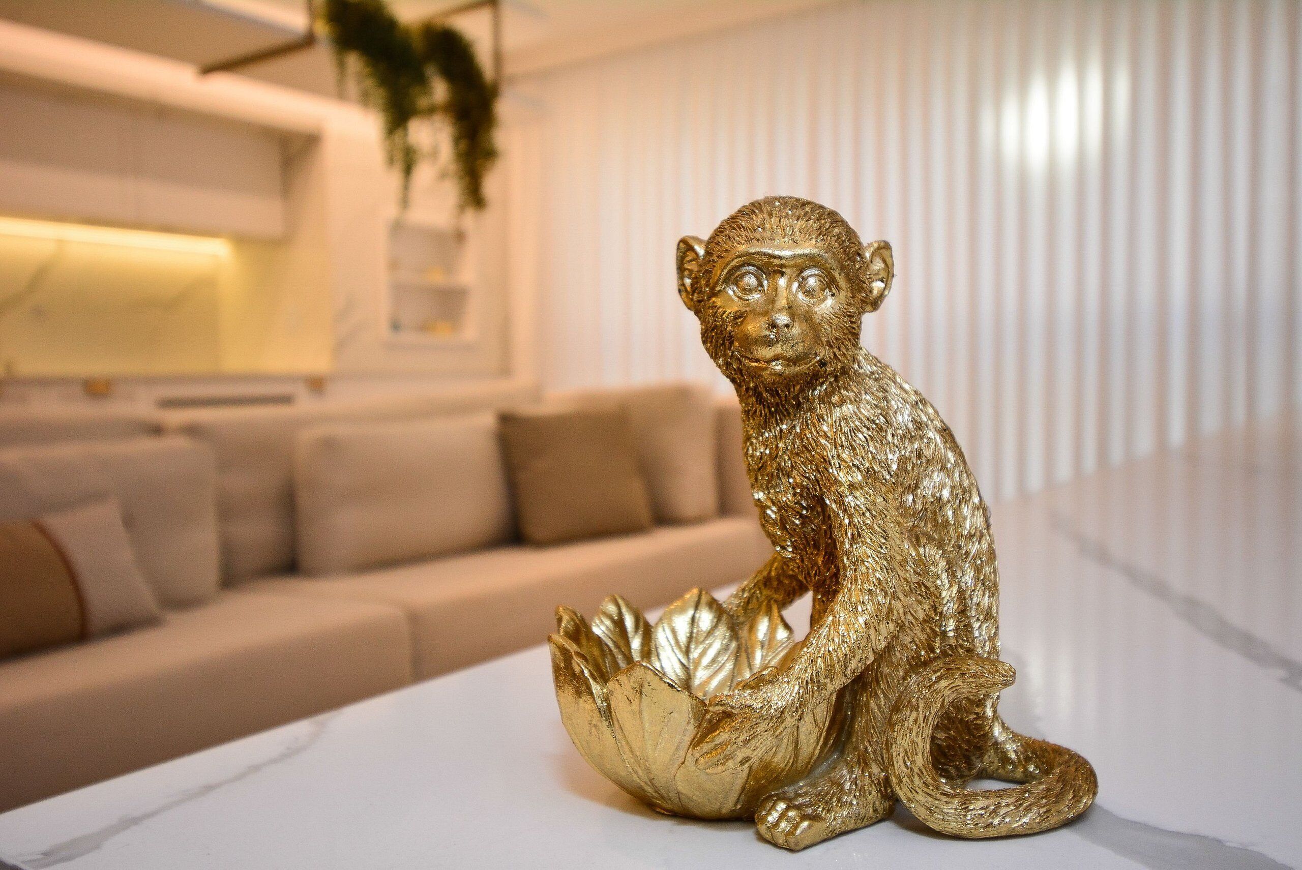 A gold monkey figurine on a white surface in a modern living room with a beige sofa and a plant in the background.