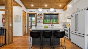 A modern kitchen with a central island, white cabinetry, and a large window offering a view of greenery. - Luxury Blackbird Cabin | Views, Hot Tub & Fire Pit (Blue Ridge)
