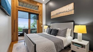 A bedroom with a large bed, two bedside lamps, a wooden headboard, a view through a glass door, and a painting on the wall. - Luxury Blackbird Cabin | Views, Hot Tub & Fire Pit (Blue Ridge)