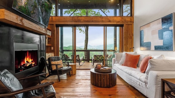 A modern living room with a fireplace, leather seating, a white sofa, and large windows offering a view of the outdoors. - Luxury Blackbird Cabin | Views, Hot Tub & Fire Pit (Blue Ridge)